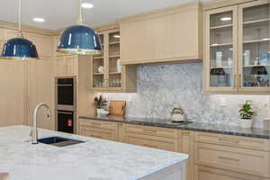 Kitchen featuring light wood finish cabinetry, glass insert cabinets, dark stone countertops, double oven, and tasteful backsplash