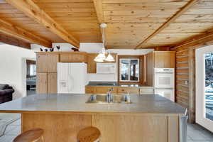 Kitchen featuring light tile patterned flooring, wooden ceiling, white appliances, light wood finish cabinetry, and a kitchen island with sink