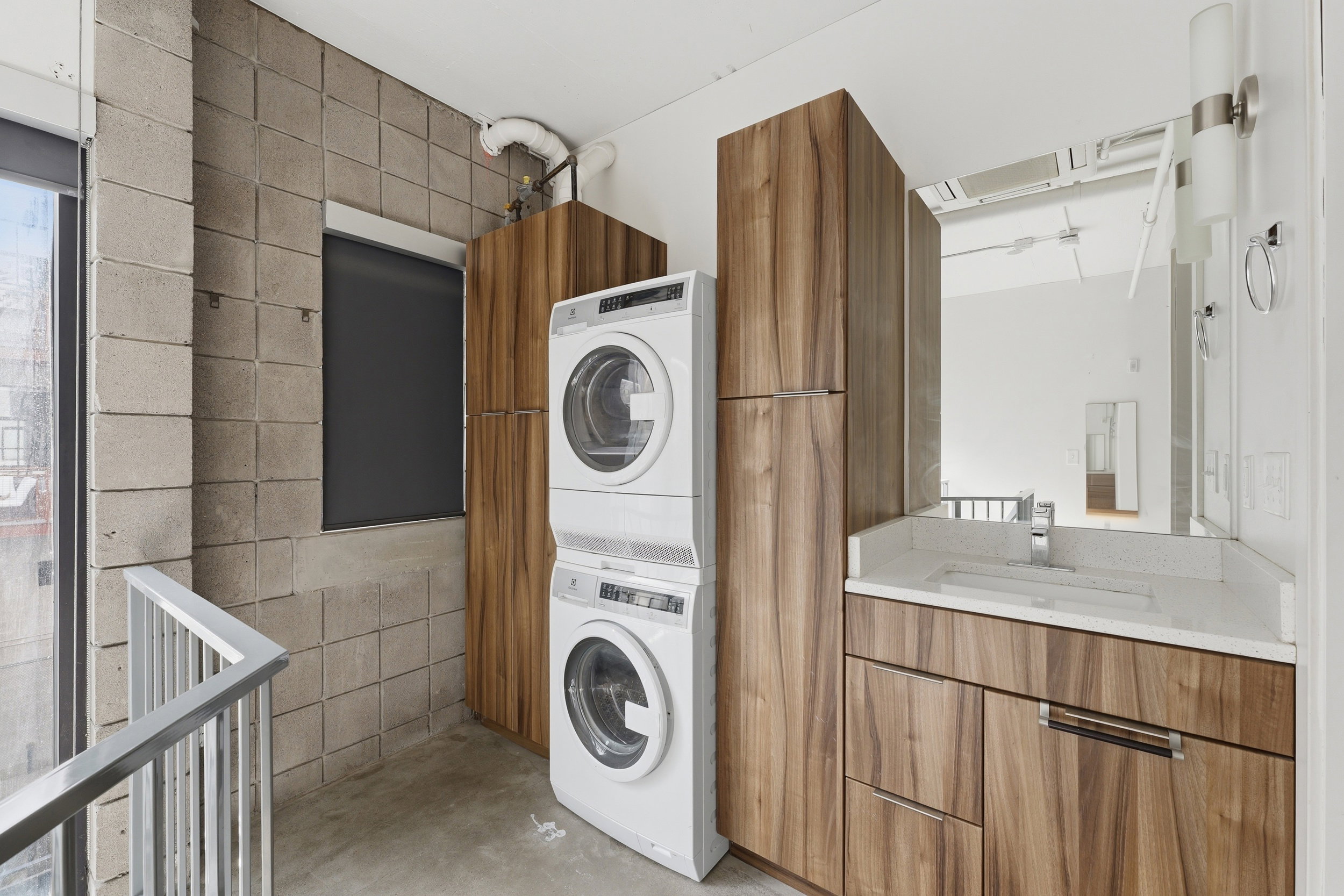 Laundry area featuring a stacked washer and dryer, wood-finish cabinetry, a single vanity with a white countertop, and exposed concrete block walls