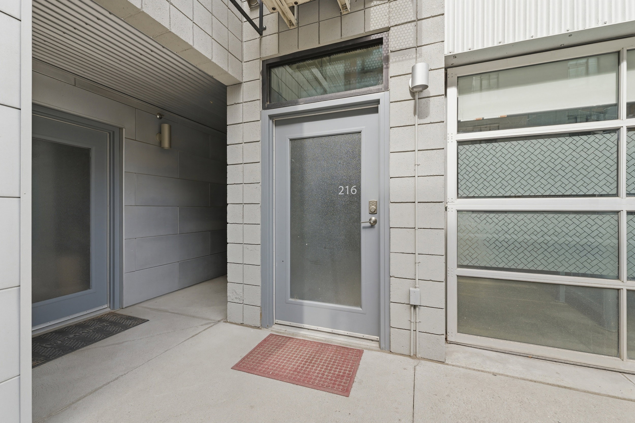 Modern residential entry with frosted glass door and transom window, featuring exterior sconce lighting, textured concrete block, and a corrugated metal awning
