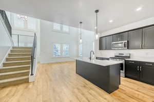 Kitchen featuring stainless steel appliances, a center island with sink, hanging light fixtures, light wood-type flooring, and light stone countertops