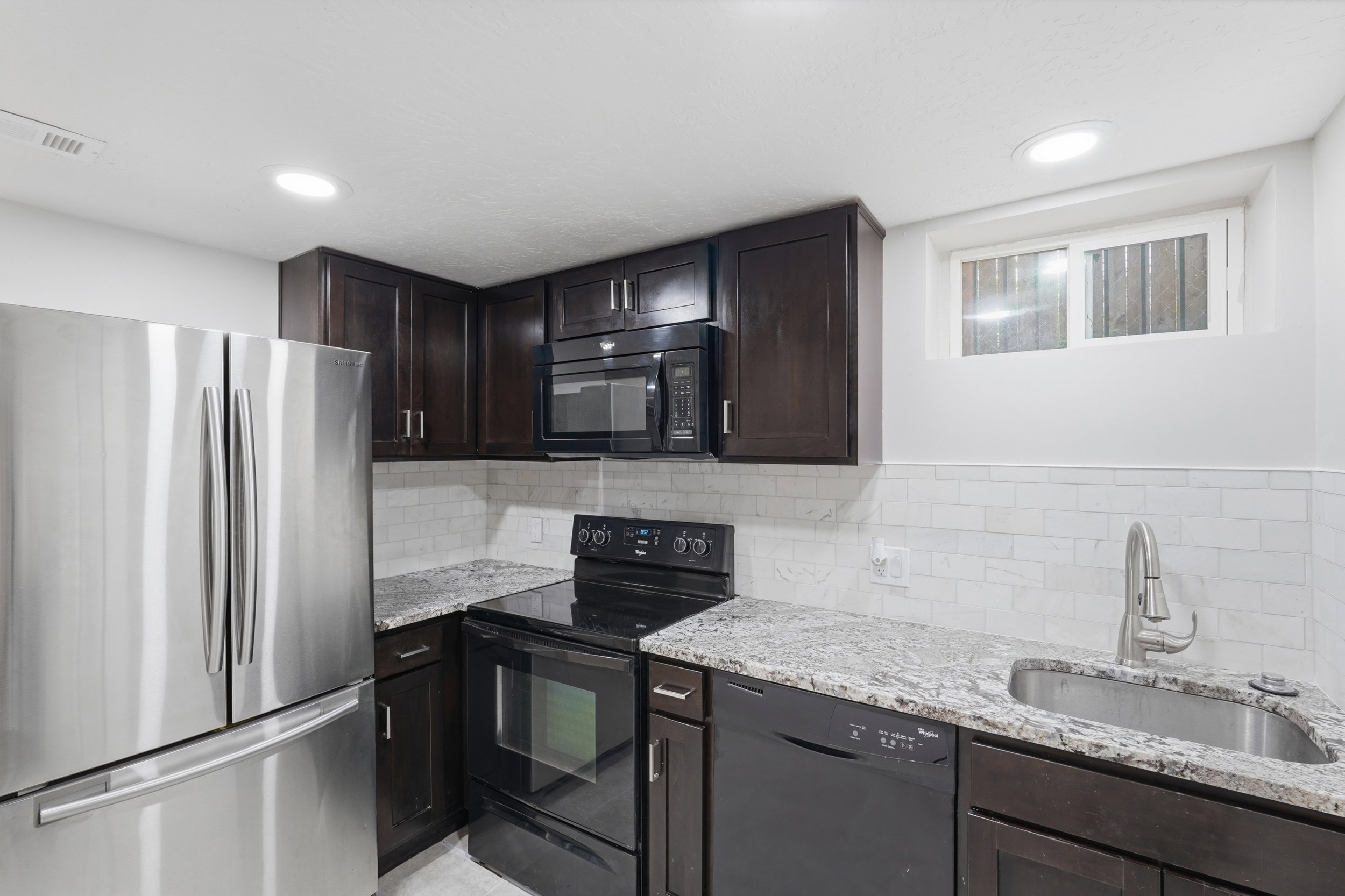 Kitchen with black appliances, dark wood finish cabinetry, light stone countertops, backsplash, and recessed lighting