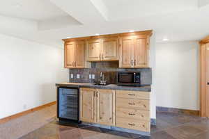 Indoor wet bar featuring light wood finish cabinetry, wine cooler, stone tile flooring, stainless steel microwave, and tasteful backsplash