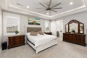 Bedroom with light colored carpet, a tray ceiling, recessed lighting, ensuite bathroom, and a ceiling fan