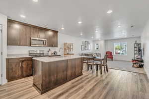 Kitchen with open floor plan, light stone countertops, stainless steel appliances, dark wood finish cabinetry, and a center island with sink