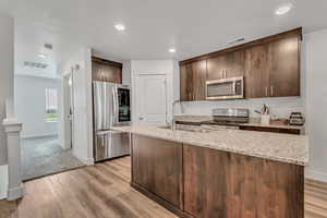 Kitchen featuring dark wood finish cabinetry, stainless steel appliances, light stone counters, light wood finished floors, and an island with sink