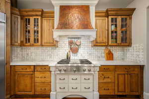 Kitchen with glass insert cabinets, light stone counters, wood finish cabinetry, stainless steel appliances, and tasteful backsplash