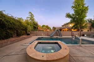 Pool at dusk featuring a water slide, a fenced backyard, a patio area, and an in-ground hot tub