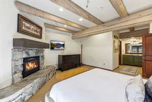 Bedroom featuring beamed ceiling, a stone fireplace, recessed lighting, ensuite bathroom, and light colored carpet