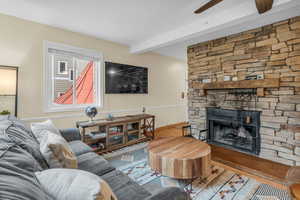 Living room with a stone fireplace, a ceiling fan, wood finished floors, and beam ceiling