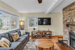 Living room featuring wood finished floors, a stone fireplace, ceiling fan, and beamed ceiling