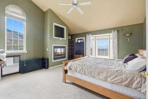 Carpeted bedroom featuring a glass covered fireplace, a ceiling fan, and vaulted ceiling