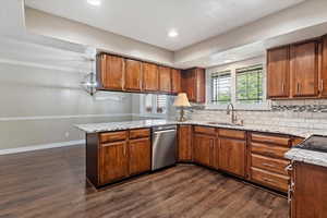 Kitchen featuring a peninsula, light stone countertops, wood finish cabinetry, stainless steel dishwasher, and dark wood-style flooring