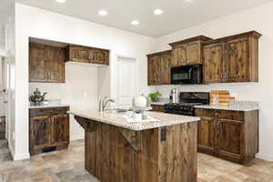 Kitchen with black appliances, an island with sink, dark wood finish cabinets, light stone countertops, and recessed lighting