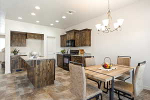 Kitchen with black appliances, dark wood finish cabinetry, a center island with sink, stone finish floors, and hanging lights