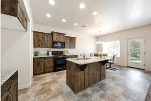 Kitchen with black appliances, a center island with sink, dark wood finish cabinets, stone finish flooring, and light stone countertops