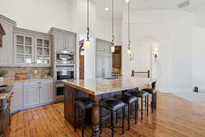 Kitchen featuring a high ceiling, glass fronted cabinets, light wood-type flooring, light stone counters, and a breakfast bar