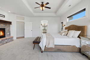 Bedroom with ensuite bathroom, a stone fireplace, ceiling fan, light carpet, and a tray ceiling