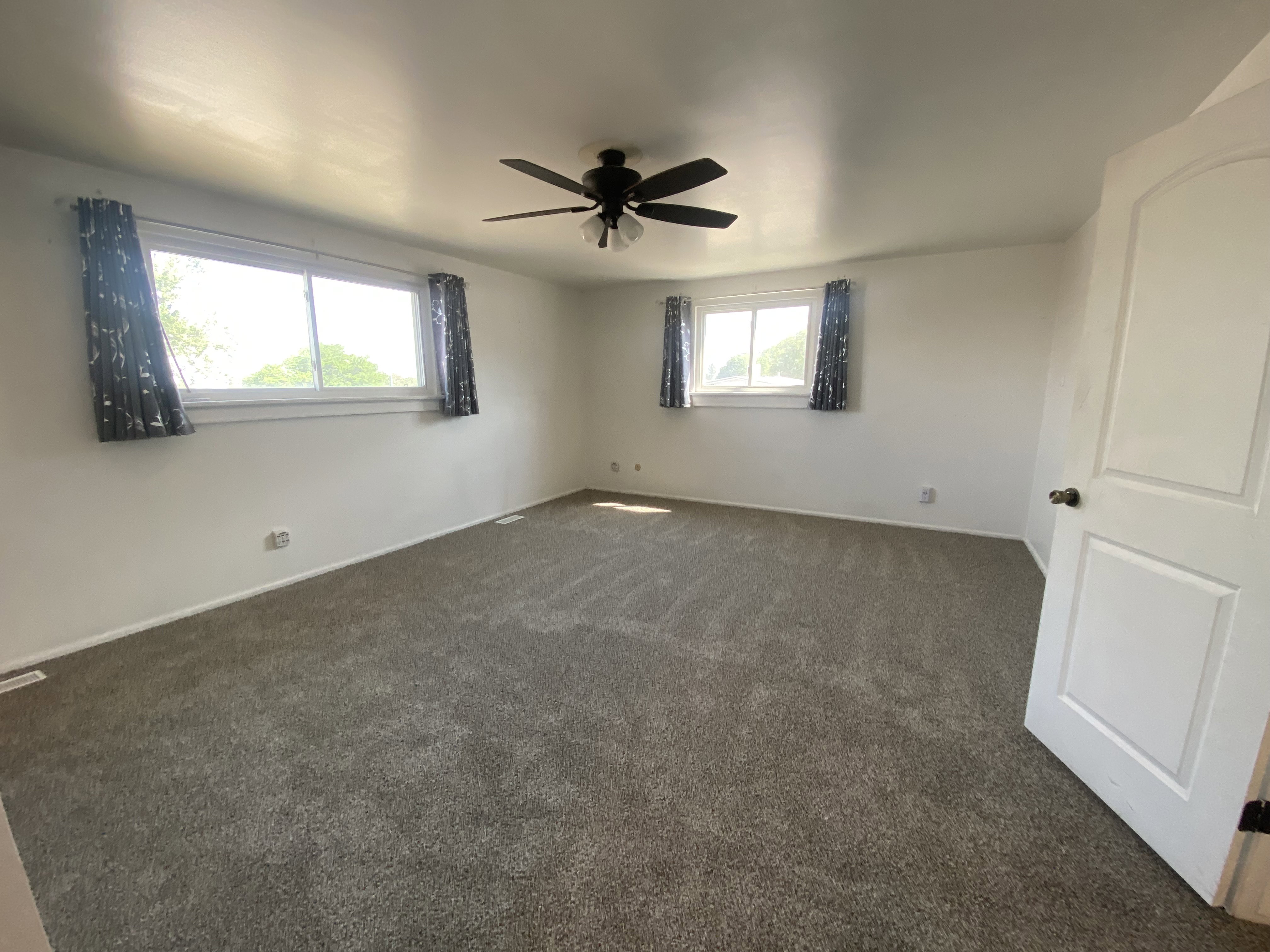 Carpeted room featuring a ceiling fan with light fixture, two windows with white trim, and a paneled interior door