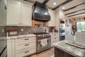 Kitchen with stainless steel appliances, light stone countertops, light wood-style flooring, beamed ceiling, and cream cabinetry