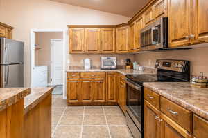 Kitchen featuring stainless steel appliances, wood finish cabinets, lofted ceiling, light tile patterned floors, and washer and clothes dryer