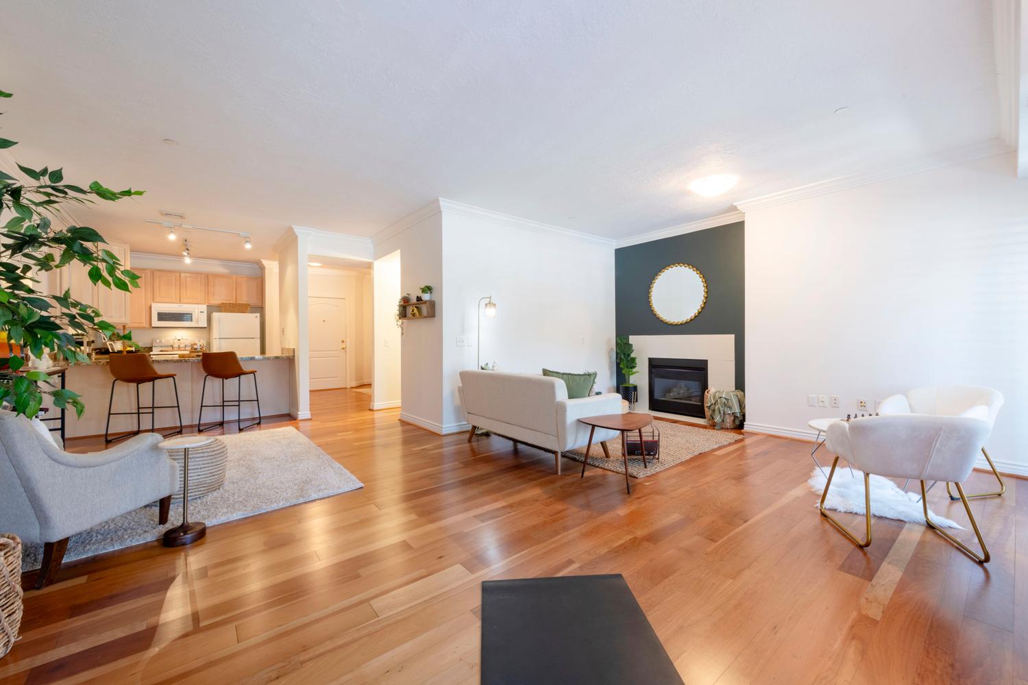 Open-concept living space featuring wood-finish flooring, a fireplace with a light-toned surround, a kitchen bar with a stone-look countertop, light wood cabinetry, and track lighting