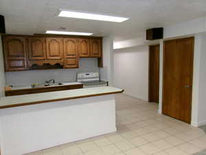 Basement Kitchen with wood finish cabinets, white electric stove, light countertops, light tile patterned floors, and a peninsula