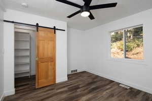 Unfurnished bedroom featuring dark wood-style flooring, a barn door, a spacious closet, ceiling fan, and a textured ceiling