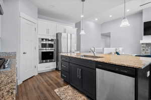 Kitchen with stainless steel appliances, two tone cabinetry, a center island with sink, light stone counters, and dark wood-type flooring
