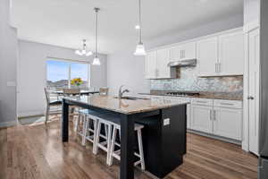 Dual tone kitchen with a breakfast bar area, light stone counters, a center island with sink, decorative backsplash, and dark wood-style floors