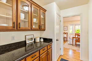 Bar area with wood finish cabinets, glass fronted cabinets, light wood-style floors, dark stone countertops, and a textured ceiling