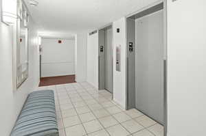 Hallway with a textured ceiling, elevator, and light tile patterned flooring