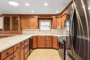 Kitchen featuring stainless steel appliances, wood finish cabinetry, glass insert cabinets, light countertops, and decorative backsplash