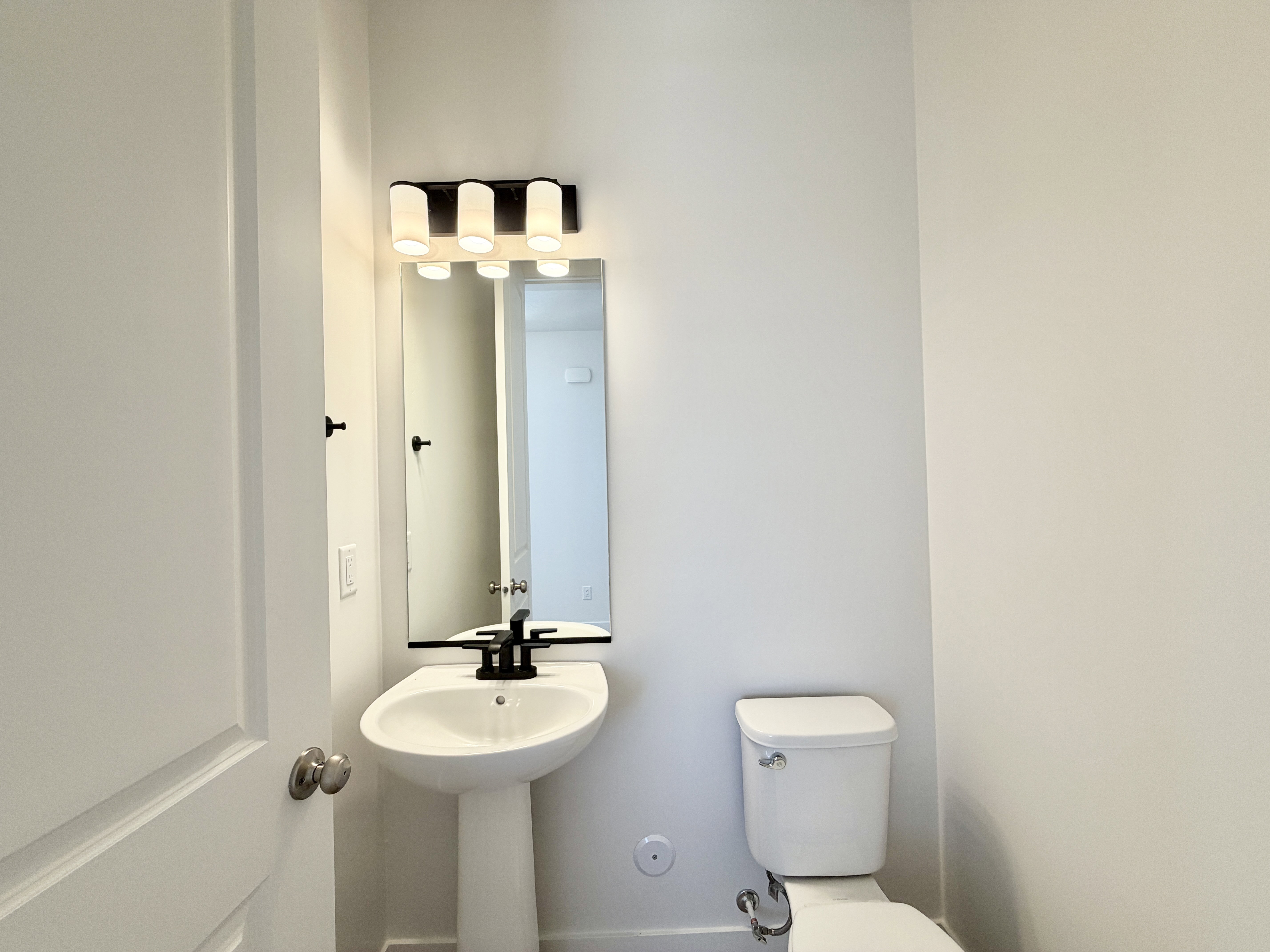 Pedestal sink with a matte black faucet, wall-mounted mirror, and a modern four-light vanity fixture