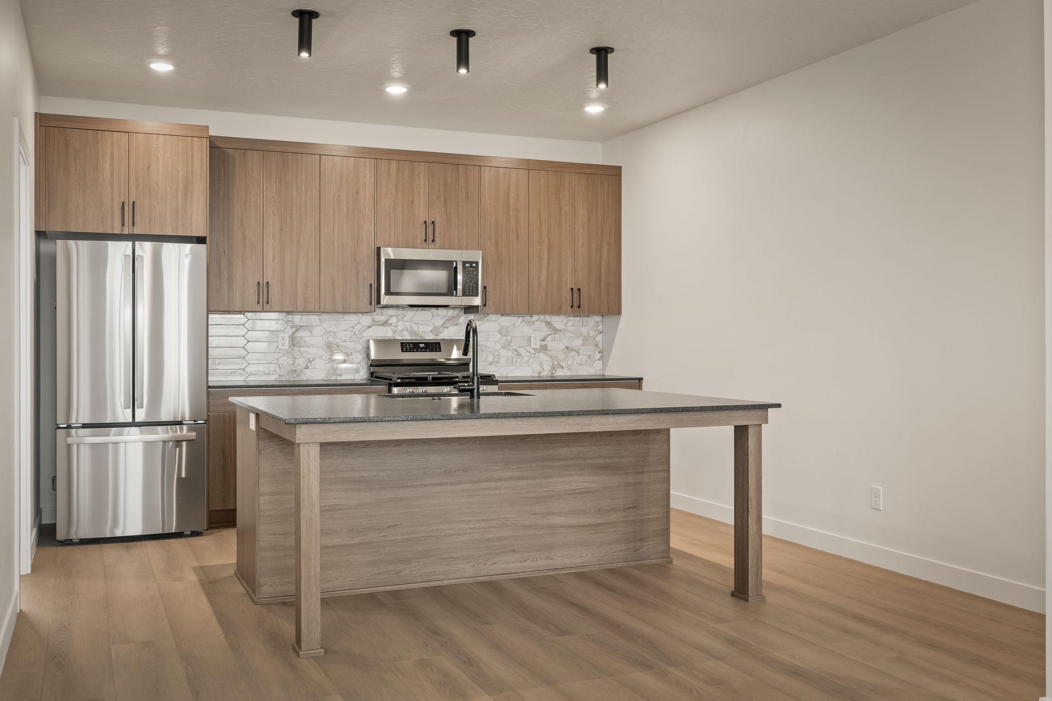 Modern kitchen featuring a spacious island with a dark countertop, stainless steel appliances, light wood-finish cabinetry, and a distinctive tile backsplash
