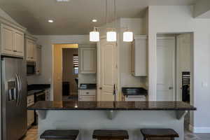 Kitchen featuring stainless steel appliances, a kitchen bar, hanging light fixtures, a kitchen island with sink, and dark stone countertops