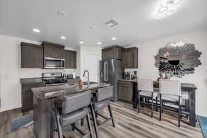 Kitchen with dark wood finish cabinets, dark stone countertops, stainless steel appliances, an island with sink, and light wood-type flooring