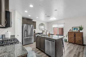 Kitchen featuring stainless steel appliances, dark stone counters, light wood-type flooring, dark wood finish cabinets, and a kitchen island with sink
