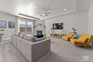 Living area with ceiling fan, a glass covered fireplace, a tray ceiling, light wood finished floors, and recessed lighting