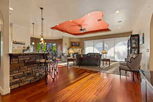 Living room with a ceiling fan, wood finished floors, a fireplace, a raised ceiling, and bar