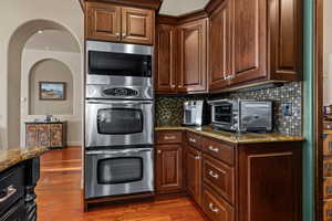 Kitchen with dark stone counters, stainless steel appliances, dark wood-style flooring, decorative backsplash, and arched walkways