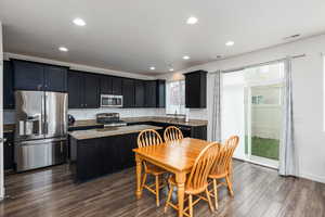 Kitchen featuring stainless steel appliances, dark cabinets, a center island, light stone countertops, and dark wood-style flooring