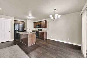 Kitchen with black appliances, dark wood-style flooring, a chandelier, dark wood finish cabinetry, and a kitchen island with sink
