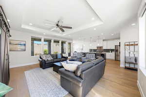 Living room with a barn door, light wood-type flooring, ceiling fan, a raised ceiling, and recessed lighting