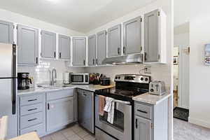 Kitchen with gray cabinetry, stainless steel appliances, light stone countertops, tasteful backsplash, and light tile patterned floors