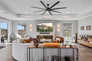 Living area with hardwood / wood-style floors, a tray ceiling, a glass covered fireplace, a ceiling fan, and recessed lighting