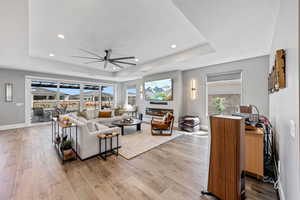 Living room featuring a tray ceiling, a ceiling fan, light wood finished floors, recessed lighting, and a glass covered fireplace