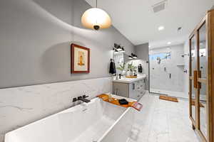 Full bathroom featuring vanity, a marble finish shower, light marble finish flooring, a soaking tub, and wainscoting