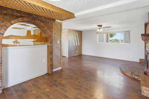 Unfurnished living room with a textured ceiling, a brick fireplace, wooden walls, dark wood finished floors, and ceiling fan