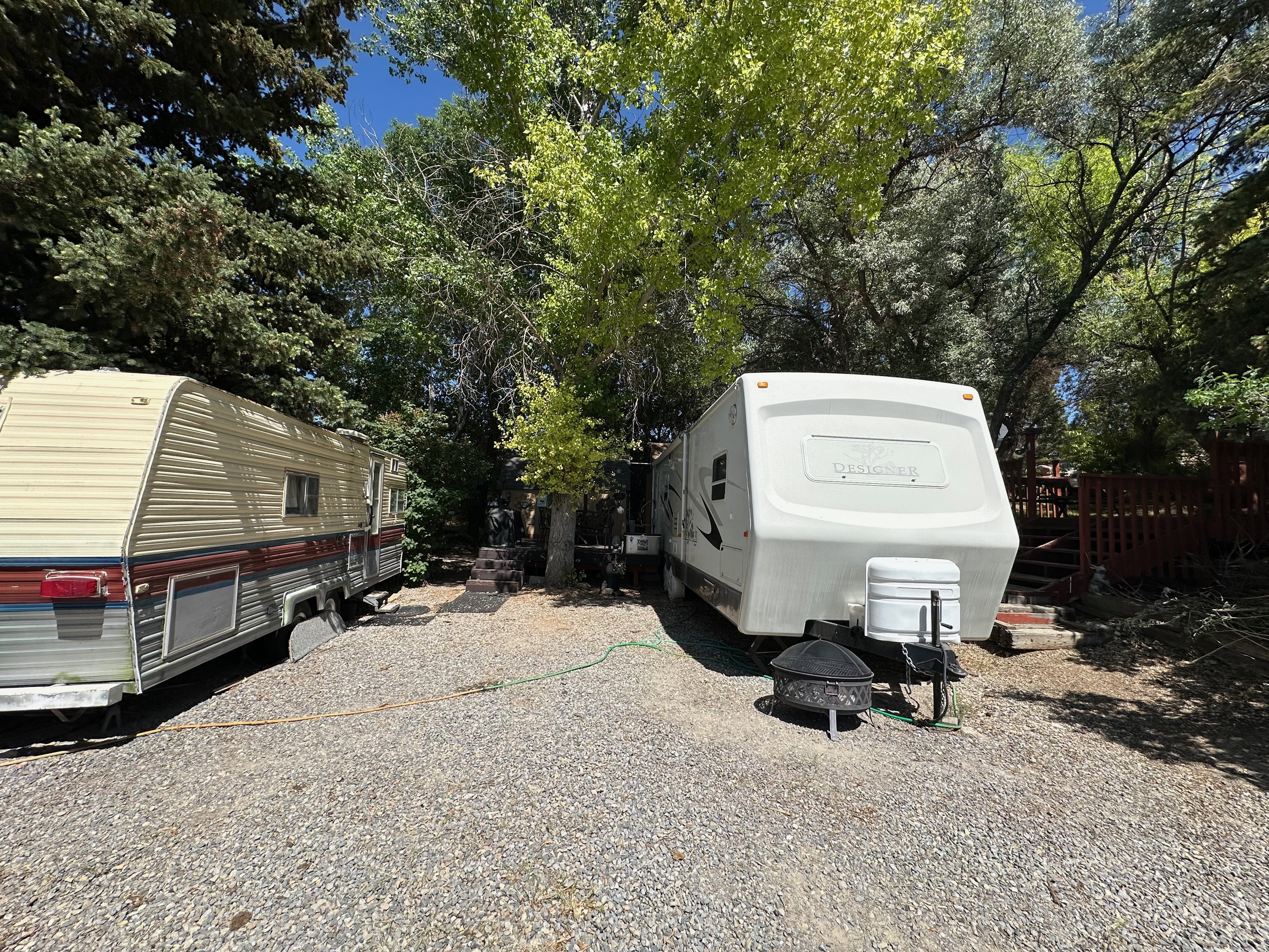 Gravel parking area featuring mature trees, a designated fire pit, and a red wooden fence